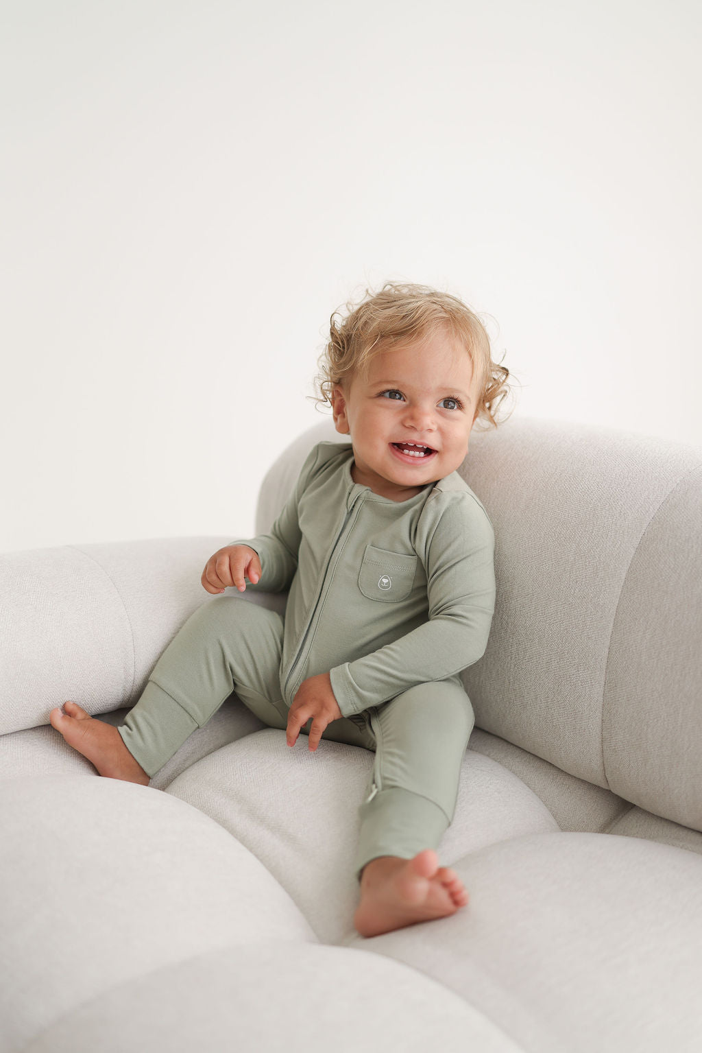 Baby in a sage green onesie sitting on a white couch