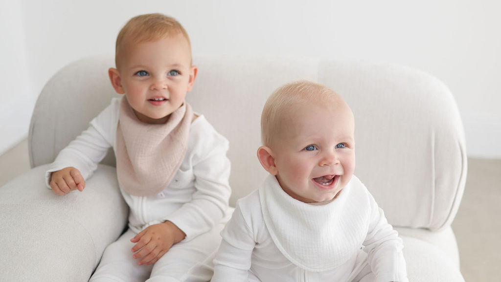 Two babies sitting on a white couch wearing light-colored clothing.