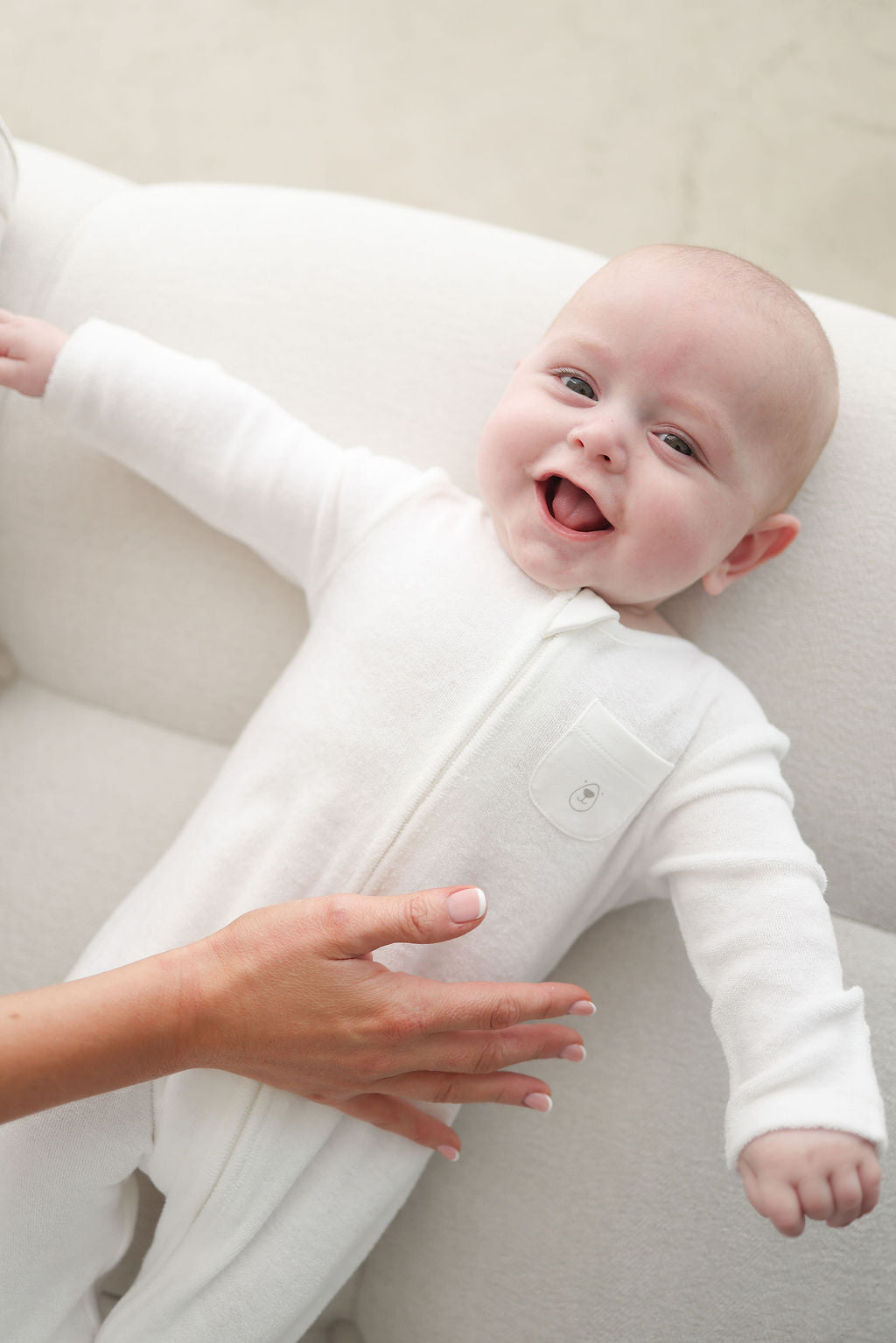 Baby in a white onesie lying on a white surface with a hand reaching out.
