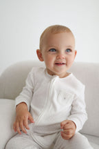 Baby wearing a white onesie sitting on a white couch.