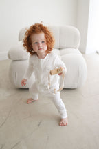 Child in white pajamas holding a toy in a minimalistic room with a white chair.